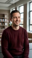 Smiling middle-aged Caucasian man in burgundy sweater sitting in modern office space with bookshelves and large windows for business portraits.