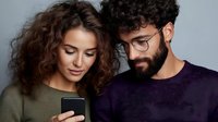 Young diverse couple looking at smartphone screen together, sharing digital content. Woman with curly hair and man with beard wearing glasses express interest.