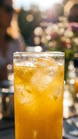 Chilled orange iced drink in a tall glass with bubbles and ice cubes, served outdoors in warm sunlight with bokeh background.