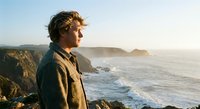 Young man with curly hair gazing at ocean waves from rocky cliff during golden hour sunset, contemplating peaceful coastal landscape scenery.