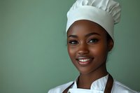 Confident young African American female chef in professional white uniform and hat smiling warmly against green background, representing culinary expertise and hospitality.