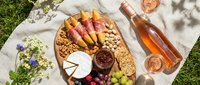 Young woman with light skin tone enjoying outdoor picnic spread with charcuterie board, wine, and fresh fruits on grass for summer entertaining and lifestyle content.