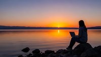 Woman working on laptop by serene lake at golden sunset with mountains in background creating peaceful remote work atmosphere.