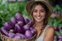 Young woman with long brown hair and fair complexion wearing a straw hat and white tank top, holding a wicker basket filled with fresh purple eggplants in a garden setting.