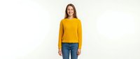 Young woman with long brown hair, Caucasian ethnicity, wearing a mustard yellow crew neck sweater and blue jeans, smiling at camera against white background. Suitable for fashion, lifestyle, and apparel marketing.