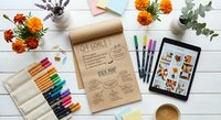 Creative workspace with colorful markers, tablet, flowers and craft supplies on white wooden desk for artistic projects and design inspiration.