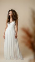 Young mixed-race woman in elegant white flowing gown with curly hair posing gracefully against warm beige background for fashion and bridal photography.