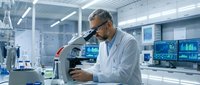 Middle-aged Caucasian male scientist in white lab coat using microscope in modern research laboratory with advanced equipment and computer monitors.