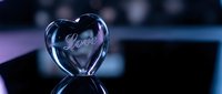 Crystal glass heart with engraved Love inscription on dark reflective surface, glowing with blue and purple light, romantic Valentine's Day concept.