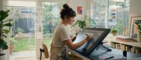 Young Asian woman digital artist working on graphics tablet in bright home studio with plants and natural light streaming through windows.