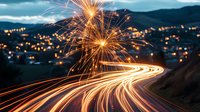 Long exposure night photography capturing light trails from vehicles on winding mountain road with bright firework sparks illuminating dark sky and city lights in background.