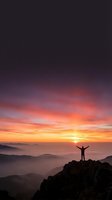Silhouette of victorious hiker with raised arms standing on mountain peak at dramatic sunrise with colorful sky and misty valleys below.