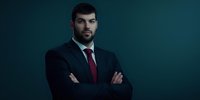 Confident middle eastern businessman in dark suit and red tie standing with arms crossed against dramatic teal background, professional corporate portrait.