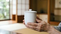 Elderly person with wrinkled hands holding white ceramic container with lid on wooden table indoors, suitable for healthcare, aging, wellness and lifestyle content.