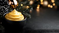 Golden creamy sauce in black bowl with festive pine branches and warm bokeh lights creating cozy holiday atmosphere for seasonal cooking and baking.