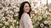 Happy young woman with dark wavy hair smiling in blooming spring garden surrounded by pink cherry blossoms and roses for lifestyle photography.