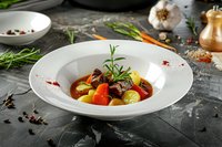 Colorful roasted vegetables with rosemary in white plate on dark background, fresh herbs and spices around. Restaurant quality food styling and presentation.