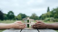 Hands protecting miniature house model on wooden table outdoors symbolizing home insurance, real estate investment and property protection concept.
