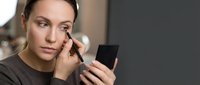 Young Caucasian woman applying makeup with brush while looking in compact mirror, beauty routine and cosmetics application in natural lighting.