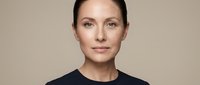 Professional middle-aged Caucasian businesswoman with natural makeup and slicked back hair wearing black top against neutral beige background for corporate branding.