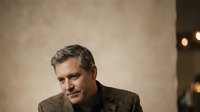 Middle-aged man with gray hair and facial stubble wearing a brown tweed jacket, looking downward in contemplative pose against neutral beige background with warm lighting.