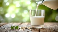 Fresh organic milk being poured into glass on rustic wooden table with natural green bokeh background for healthy breakfast and dairy products.
