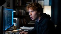 Young male programmer with reddish-brown hair, Caucasian ethnicity, wearing dark clothing, focused on computer code displayed on monitor in modern office workspace at night.