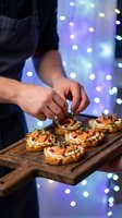 Male chef hands garnishing gourmet appetizers on wooden board with festive bokeh lights background for holiday catering and fine dining presentation.