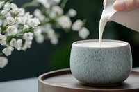 Pouring fresh milk into minimalist ceramic bowl on wooden tray with white flowers in soft focus background, creating serene morning atmosphere.