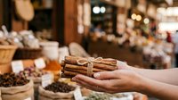 Closeup of hand holding cinnamon sticks bundled with twine in spice market with blurred vendor stall background, suitable for food, culinary, and retail commerce projects.