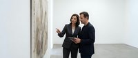 Professional Asian woman and Caucasian man in business suits discussing artwork in modern gallery space with contemporary painting on white wall.