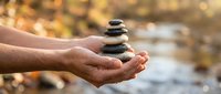 Hands gently holding balanced zen stones in cupped palms with soft natural bokeh background for meditation wellness and mindfulness concepts.