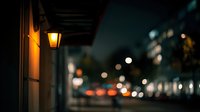 Vintage street lamp illuminating building entrance at night with blurred city lights and traffic in background creating atmospheric urban scene.