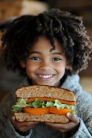 Happy African American child with curly hair holding healthy vegetable sandwich with tomatoes and lettuce on whole grain bread for nutritious meal concept.