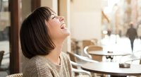 Happy Asian woman laughing joyfully at outdoor cafe terrace during sunny day, expressing genuine happiness and positive emotions in urban setting.