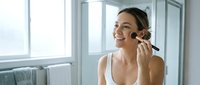 Young Caucasian woman applying makeup with brush in bright modern bathroom, morning beauty routine skincare cosmetics preparation getting ready.