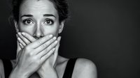 Young woman with hands covering mouth expressing shock or fear against dark background, dramatic black and white portrait conveying emotional distress.