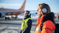 Female airport ground crew member in orange safety jacket and headset communicating on airfield with aircraft and colleague in background during daytime operations.