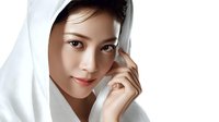 Young Asian woman with natural makeup wearing white fabric draped over head, looking at camera with gentle smile against light background. Soft beauty portrait.