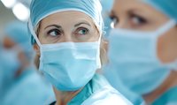 Female medical professional in surgical attire and protective face mask looking confidently at camera, with colleague in background during hospital procedure.