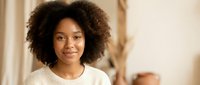 Young African American woman with curly afro hair smiling warmly in cozy indoor setting wearing white sweater for lifestyle and portrait photography.