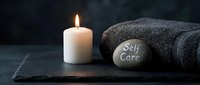 Spa wellness setup with lit candle, folded towel, and stone engraved with Self Care on dark slate background for relaxation themes.