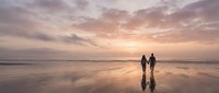 Couple walking hand in hand on wet sand beach at sunset with dramatic pink and orange sky reflecting on water creating romantic peaceful moment.