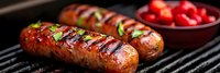 Grilled sausages with herbs on barbecue grill next to bowl of cherry tomatoes for summer cooking and outdoor dining.