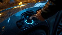 Electric vehicle charging port glowing blue in dark rainy conditions, close-up of hand plugging in charger with water droplets on car surface.