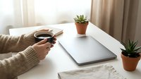 Person in beige sweater holding coffee cup at white desk with closed laptop and small potted succulents in terracotta pots creating cozy home office workspace.