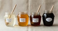 Four glass jars filled with different honey varieties from light to dark, each with wooden dipper and handwritten label on neutral linen background.