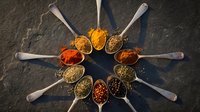 Colorful spices and herbs arranged in metal spoons forming circular pattern on dark slate background for cooking and culinary seasoning applications.