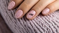Trendy manicure featuring pink polka dot nails with love text on ring finger against cozy knitted sweater background, showcasing modern nail art design and feminine style.