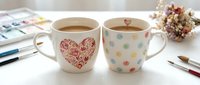 Two ceramic mugs with hot beverages on white desk with art supplies and dried flowers decoration.